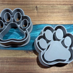 Puede incluir: Dos cortadores de galletas grises con forma de huella de pata. Los cortadores son de pl&aacute;stico y tienen un dise&ntilde;o detallado. Las huellas de pata est&aacute;n sobre una superficie de madera con una incrustaci&oacute;n de resina epoxi azul y verde.