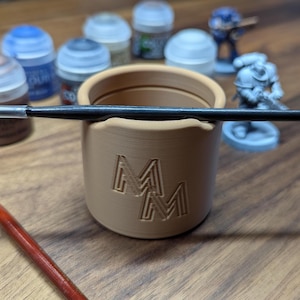Puede incluir: Un portacepillos cil&iacute;ndrico beige con las letras "MM" grabadas en el lateral. Un pincel negro descansa sobre el borde. Figuras en miniatura y botes de pintura en el fondo, sugieren un tema de afici&oacute;n o manualidades.