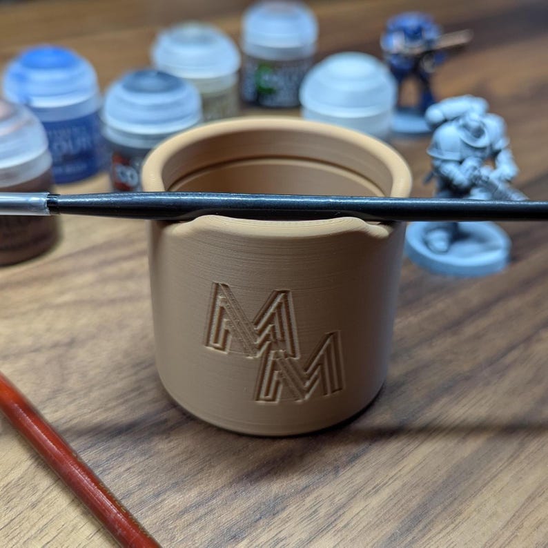 Puede incluir: Un portacepillos cil&iacute;ndrico beige con las letras "MM" grabadas en el lateral. Un pincel negro descansa sobre el borde. Figuras en miniatura y botes de pintura en el fondo, sugieren un tema de afici&oacute;n o manualidades.