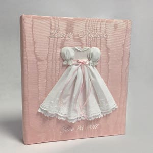 May include: A light pink fabric-covered memory book featuring a white dress appliqu&eacute; with puff sleeves and a pink ribbon tie. The name Lucy Marie is embroidered in white above the dress, and the date June 26, 2017 is embroidered in white below.