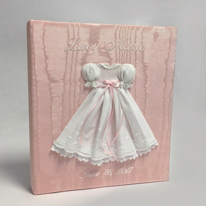 May include: A light pink fabric-covered memory book featuring a white dress appliqu&eacute; with puff sleeves and a pink ribbon tie. The name Lucy Marie is embroidered in white above the dress, and the date June 26, 2017 is embroidered in white below.