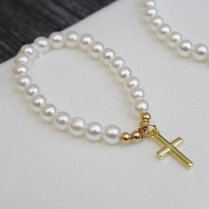 May include: A white pearl bracelet with a gold cross charm. The bracelet is made of small, round pearls and has a simple, elegant design.