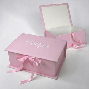 May include: Two rectangular light pink gift boxes with hinged lids. One box is closed and features the name Cooper embroidered in white script on the lid, secured with a pink ribbon. The second box is open, revealing a white interior lining.