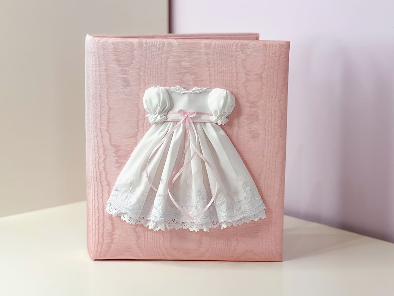 May include: A light pink photo album with a textured cover featuring a white dress appliqu&eacute;. The dress has puffed sleeves, a pink ribbon tied in a bow at the waist, and a white lace hem with intricate embroidery details across the skirt.
