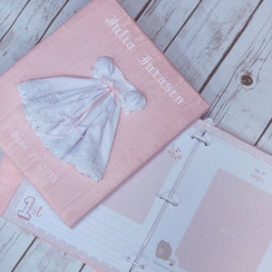 May include: A light pink fabric-covered photo album featuring the embroidered name Julia Juraszy and the date June 27 2018. A small white dress with lace trim and a pink ribbon is attached to the cover. The open album shows pages with the text 1st and my 1st steps.