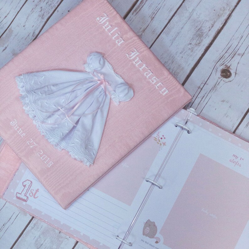 May include: A light pink fabric-covered photo album featuring the embroidered name Julia Juraszy and the date June 27 2018. A small white dress with lace trim and a pink ribbon is attached to the cover. The open album shows pages with the text 1st and my 1st steps.