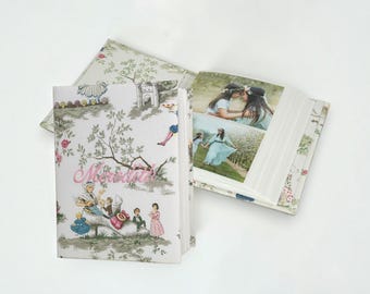 May include: A photo album with a fabric cover featuring a toile-style print of figures and trees in muted green and grey tones. The name Meredith is embroidered in pink script on the front. The open album displays two color photographs of people.