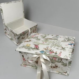 May include: A rectangular keepsake box with a hinged lid, covered in a white fabric featuring a colorful toile-style print of figures and animals. The lid is tied with a white ribbon and features the embroidered text Baby Keepsake.