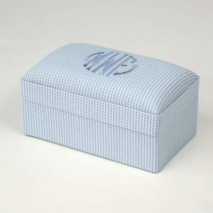 May include: A rectangular box with a light blue gingham pattern and a curved lid. The lid is adorned with a darker blue embroidered monogram. The box is designed for storage or decorative purposes, with a classic aesthetic.