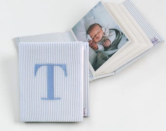 May include: A fabric-covered photo album featuring a light blue and white pinstripe pattern. The front cover is embroidered with a large, centered blue letter T. The album is open, displaying a photograph of a newborn baby holding a small toy.
