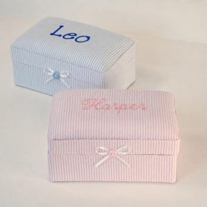 Könnte beinhalten: Zwei rechteckige, stoffbezogene Aufbewahrungsboxen mit Klappdeckeln. Eine Box hat blau-weiße Streifen mit dem in Blau gestickten Namen Leo und einer blauen Knopfschleife. Die andere Box hat rosa-weiße Streifen mit dem in Rosa gestickten Namen Harper und einer rosa Knopfschleife.