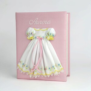 May include: A light pink photo album featuring the embroidered name Aurora in white script. Attached to the cover is a white fabric dress with puffed sleeves, yellow trim, pink ribbon ties, and a floral embroidered hem with white lace.