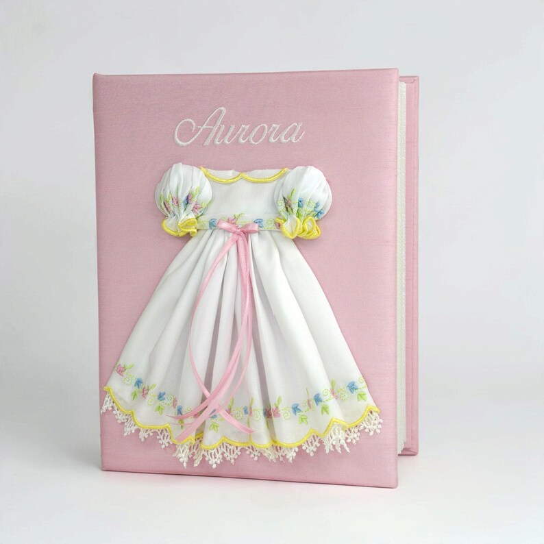 May include: A light pink photo album featuring the embroidered name Aurora in white script. Attached to the cover is a white fabric dress with puffed sleeves, yellow trim, pink ribbon ties, and a floral embroidered hem with white lace.