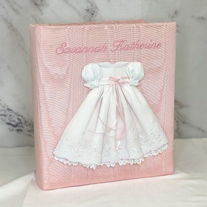 May include: A light pink photo album with a textured finish featuring the embroidered name Savannah Katherine in pink thread. A white dress with puff sleeves, a pink ribbon bow at the waist, and intricate lace trim at the hem is attached.