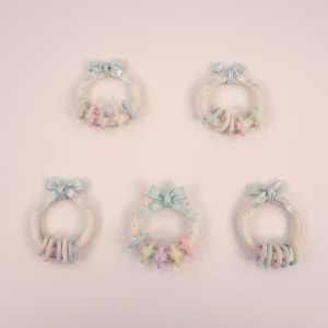 Puede incluir: Cinco coronas de color pastel con lunares azules y lazos azules. Cada corona tiene un juego de anillos de color pastel colgando de ella.