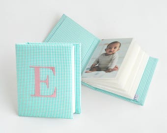 May include: A fabric photo album with a teal and white gingham pattern. The front cover features a large, embroidered pink letter E. The album is open, revealing a photograph of a baby wearing a long-sleeved shirt inside the clear plastic sleeves.