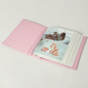 May include: An open photo album with a solid light pink fabric cover. The interior pages display two photographs: one showing a person holding a baby, and another showing a baby lying down wearing a white outfit with a pink bow headband.