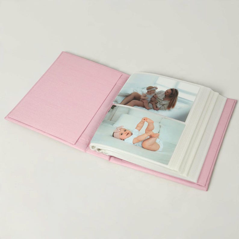 May include: An open photo album with a solid light pink fabric cover. The interior pages display two photographs: one showing a person holding a baby, and another showing a baby lying down wearing a white outfit with a pink bow headband.
