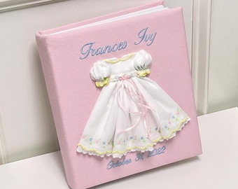 May include: A light pink photo album featuring an embroidered white dress with puff sleeves and ribbon accents. The cover displays the name Frances Ivy in blue script embroidery above the dress and October 31, 2022 in blue below.