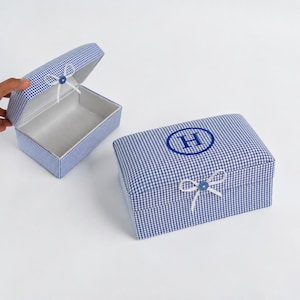 Könnte beinhalten: Zwei rechteckige Aufbewahrungsboxen, die mit blau-weißem Vichy-Stoff bezogen sind. Eine Box ist geöffnet und zeigt ein weißes Inneres, während die andere geschlossen ist und ein blau gesticktes H in einem Kreis sowie eine kleine weiße Schleife aufweist.