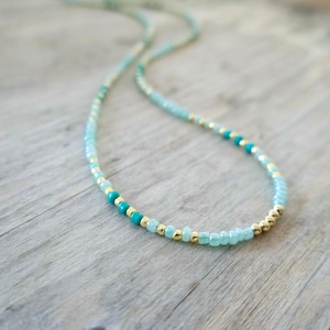May include: Delicate beaded necklace featuring small, light blue, turquoise, and gold-colored beads. The necklace is strung on a thin, flexible cord, creating a subtle and elegant accessory. The beads are arranged in a repeating pattern.