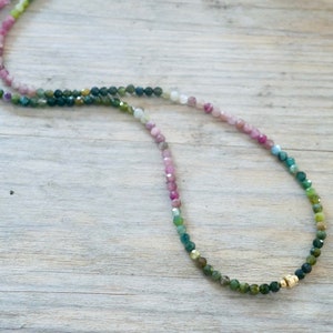 May include: A multi-colored gemstone necklace with a gradient of pink, blue, and green beads. The necklace is made with small, faceted beads and has a gold clasp.