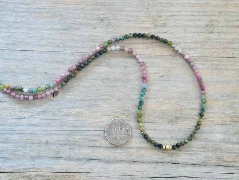 May include: A multi-colored gemstone necklace with a gold bead accent. The necklace is made of small, round beads in shades of pink, green, and blue.