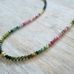 May include: A multi-colored gemstone necklace with a gradient of pink, green, and black beads. The necklace is made with small, round beads and has a gold clasp.