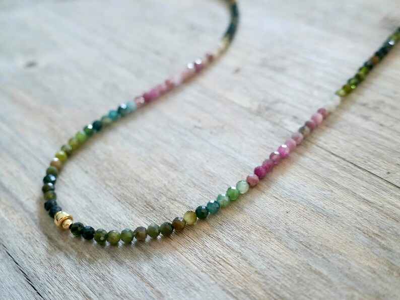 May include: A multi-colored gemstone necklace with a gradient of pink, green, and black beads. The necklace is made with small, round beads and has a gold clasp.