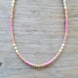 May include: A delicate beaded necklace featuring a pattern of pink, white, and gold beads. The necklace has a simple design with a strand of small, round beads in alternating colors. The necklace is displayed on a wooden surface.