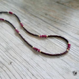 May include: A delicate necklace featuring small, dark brown beads interspersed with larger, vibrant red gemstones and tiny gold-colored accents. The necklace is displayed on a light brown wooden surface.
