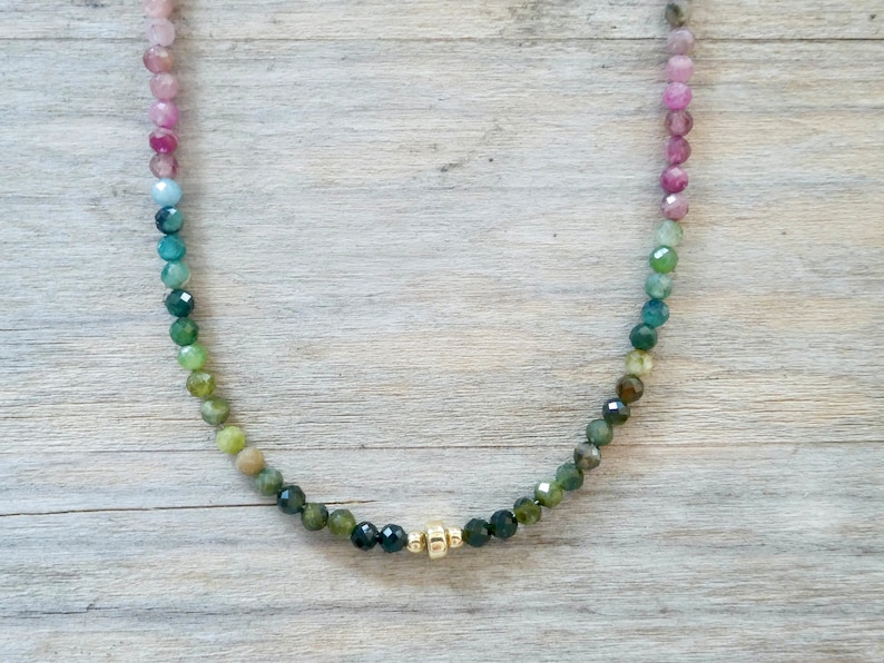 May include: A delicate necklace made with faceted gemstone beads in a gradient of pink, blue, green, and black. The necklace features a gold accent in the center.