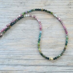 May include: A multi-colored gemstone necklace with a gold clasp. The necklace is made of small, round beads that are pink, blue, green, and black.