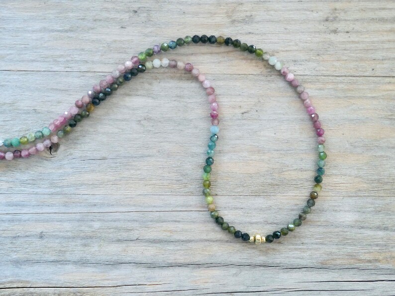 May include: A multi-colored gemstone necklace with a gold clasp. The necklace is made of small, round beads that are pink, blue, green, and black.