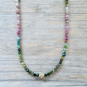 May include: A rainbow tourmaline gemstone necklace with a gold clasp. The necklace is made of small, round beads that are strung together in a gradient of colors, from pink to green.