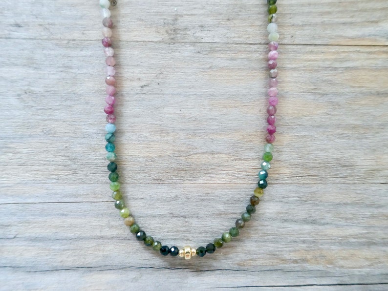May include: A rainbow tourmaline gemstone necklace with a gold clasp. The necklace is made of small, round beads that are strung together in a gradient of colors, from pink to green.
