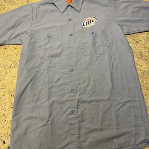 May include: A light blue work shirt with a short sleeve and a Miller Lite logo embroidered on the left chest.