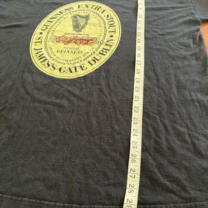 Puede incluir: Camiseta negra con un gr&aacute;fico circular dorado y rojo que presenta el texto "St. James's Gate Dublin" y "Guinness Extra Stout" con un arpa y las palabras "Brewers of Dublin Since 1778".