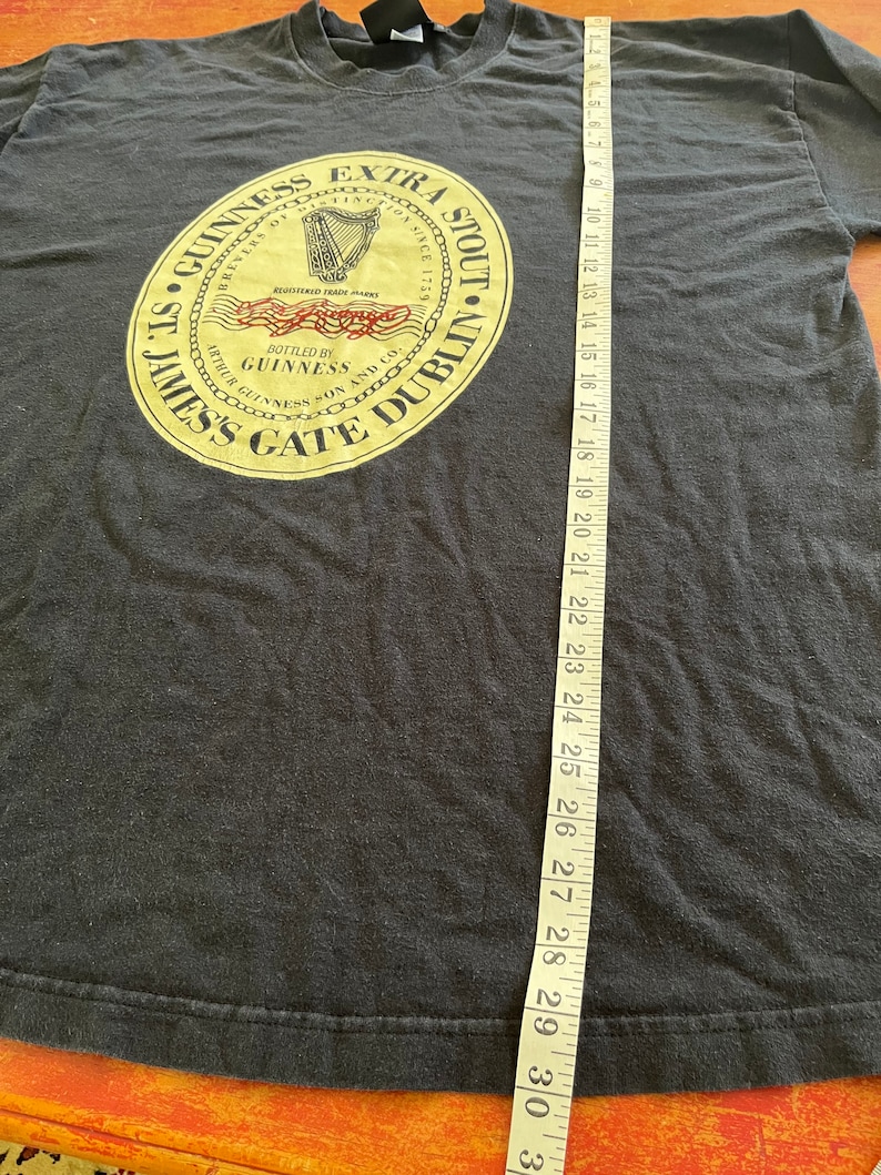 Puede incluir: Camiseta negra con un gr&aacute;fico circular dorado y rojo que presenta el texto "St. James's Gate Dublin" y "Guinness Extra Stout" con un arpa y las palabras "Brewers of Dublin Since 1778".