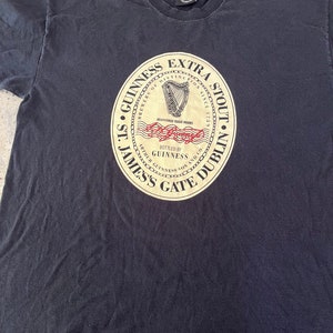 Puede incluir: Camiseta negra con un gr&aacute;fico circular de estilo vintage del logotipo de Guinness Extra Stout. El logotipo presenta un arpa dorada, las palabras "Brewers of Distinction Since 1778", "St. James's Gate Dublin" y "Bottled by Guinness".