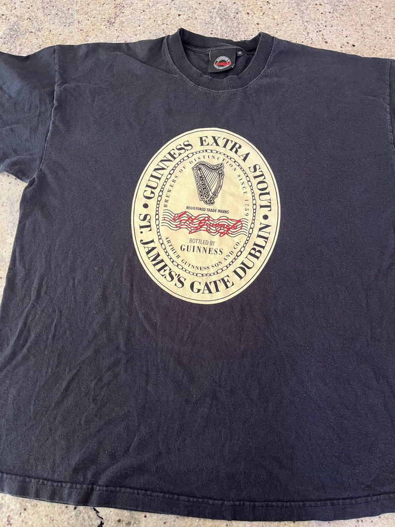 Puede incluir: Camiseta negra con un gr&aacute;fico circular de estilo vintage del logotipo de Guinness Extra Stout. El logotipo presenta un arpa dorada, las palabras "Brewers of Distinction Since 1778", "St. James's Gate Dublin" y "Bottled by Guinness".
