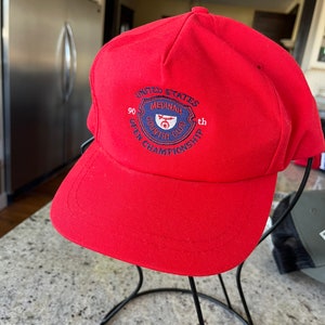 Op de afbeelding: Rode baseballpet met een donkerblauwe geborduurde patch met de tekst "United States 90th Open Championship Medinah Country Club".
