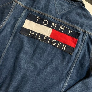 May include: A blue denim jacket with a large Tommy Hilfiger logo patch on the back. The patch is red, white, and blue and features the brand name in white lettering.