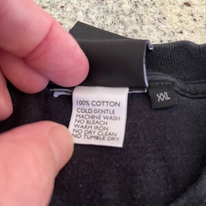Puede incluir: Una camiseta de algod&oacute;n negro con una etiqueta de cuidado blanca que dice "100% Algod&oacute;n, Lavado a m&aacute;quina en fr&iacute;o delicado, Sin blanqueador, Planchado en caliente, No lavar en seco, No secar en secadora" y una etiqueta negra que dice "XXL".
