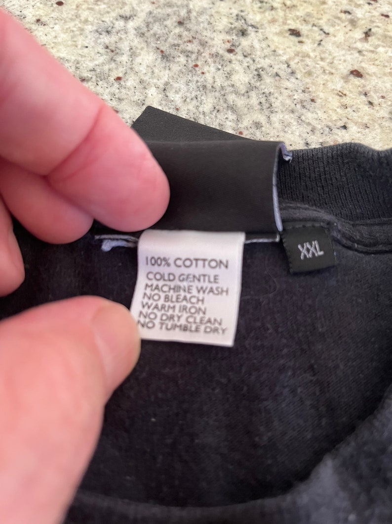 Puede incluir: Una camiseta de algod&oacute;n negro con una etiqueta de cuidado blanca que dice "100% Algod&oacute;n, Lavado a m&aacute;quina en fr&iacute;o delicado, Sin blanqueador, Planchado en caliente, No lavar en seco, No secar en secadora" y una etiqueta negra que dice "XXL".