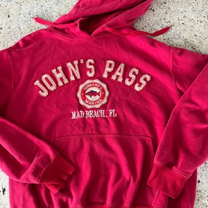 May include: Red hooded sweatshirt with the text "John's Pass" in white with a white embroidered logo of a fish and the text "Hubbard's Marina" and "Mad Beach, FL" below.