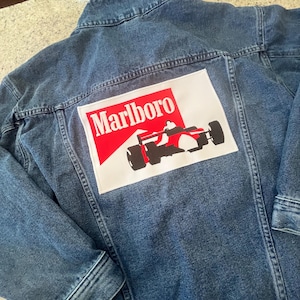 May include: A blue denim jacket featuring a large rectangular patch on the back. The patch displays the red and white Marlboro logo with a black and red graphic of a Formula One race car. The jacket has a standard collar and long sleeves.
