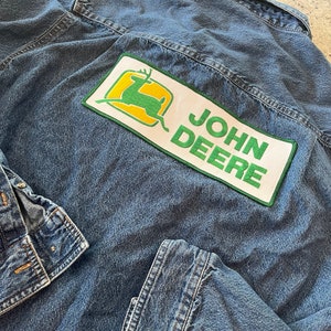 Op de afbeelding: Een blauwe denimjas met een groene en gele John Deere-patch op de rug. De patch toont een gestileerd hert met geweien.