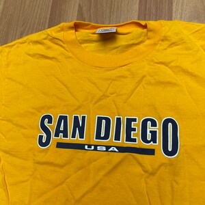Peut inclure: T-shirt jaune avec un motif noir et blanc qui dit "San Diego USA" en caractères gras.