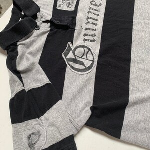 May include: A gray and black striped polo shirt with a black embroidered logo on the left chest and a black embroidered patch on the right sleeve. The logo says "Unlimited Sacrifice".